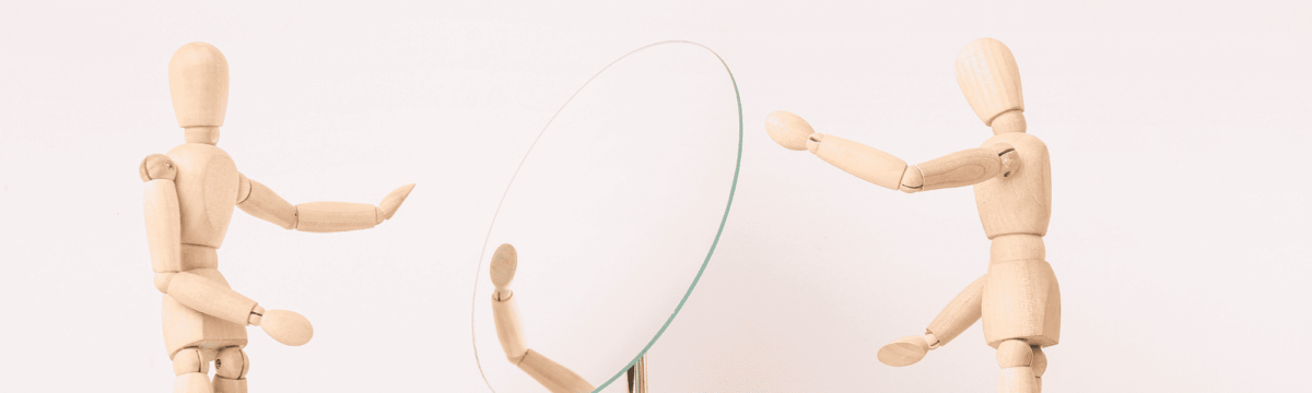 Two wooden figures, one reaching out to another who is focused on their reflection in a mirror