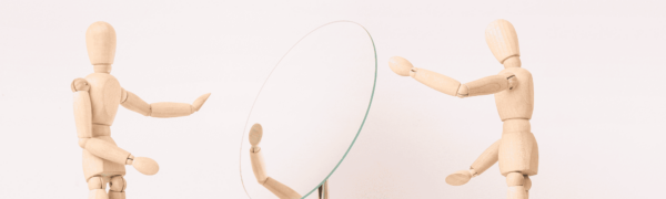 Two wooden figures, one reaching out to another who is focused on their reflection in a mirror